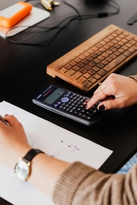 A person uses a calculator to determine fair value of a business. A wood keyboard and notebook are also on the desktop.